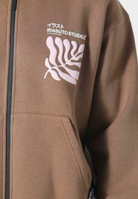 Braune Zip-Hoodie aus weichem Stoff, mit einem weißen Grafikdesign von Blättern und dem Markenlogo auf der Brust sowie vorderen Taschen.