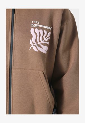 Ruskea vetoketjuhuppari, joka on valmistettu pehmeästä kankaasta. Hupparissa on rinnan kohdalla valkoinen graafinen lehtimuotoilu ja brändilogo, sekä etutaskut.