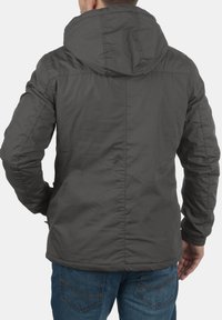 Grijze hoodie met een textuurafwerking, zichtbare naden en elastische boorden. Draagt over blauwe Jeans en toont een eenvoudig, functioneel ontwerp.