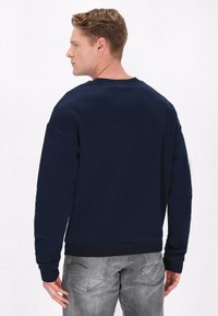 Marineblauer Sweatshirt mit Rundhalsausschnitt, breiten Schultern und gerippten Bündchen, kombiniert mit grauen Denim-Hosen, von hinten betrachtet.