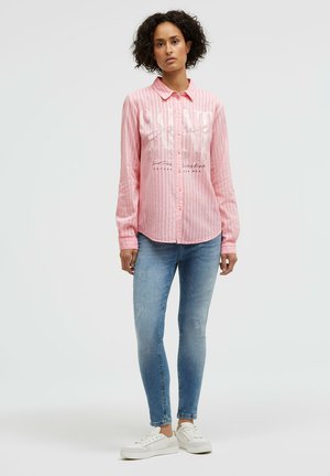 Frau steht vor einem schlichten Hintergrund, trägt ein pink gestreiftes Hemd mit Knöpfen, hellblaue Jeans und weiße Turnschuhe.