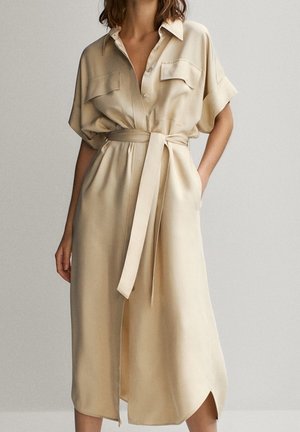 Beige midi-pituinen paitamekko, jossa on lyhyet hihat, rintataskut, kaulus, napitus edessä ja vyötärölle sidottava vyö.