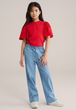 Piros pamut póló szemes díszítéssel a pöttyös ujjakon, világoskék, függőleges csíkos, bő fazonú nadrággal és fehér sportcipőkkel párosítva.