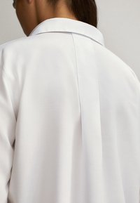 Valkoinen, sileä kangaspaita klassisella kauluksessa. Takana on keskisauma ja hienovarainen tekstuuri. Yksinkertainen, koristelematon muotoilu.