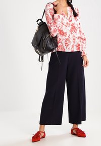 Blusa bianca con motivo floreale rosso, pantaloni larghi blu navy, scarpe rosse con frange e uno zaino in pelle nera. Outfit casual con colori vivaci.