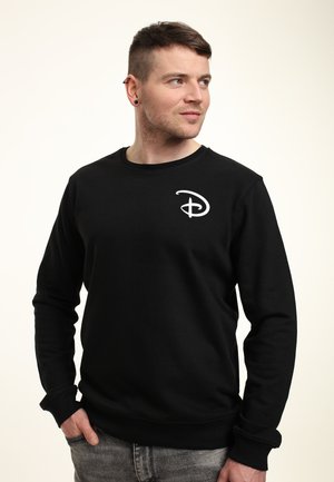 Mężczyzna o krótkich brązowych włosach, ubrany w czarną bluzę z białym logo Disneya, stoi z jedną ręką w kieszeni, patrząc w prawo na jednolitym tle.