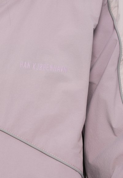 Ανοιχτό μωβ μπουφάν με ματ υφή, που διαθέτει λευκή ραφή και το όνομα της μάρκας "HAN KJØBENHAVN" σε ελαφρύτερη απόχρωση μωβ.