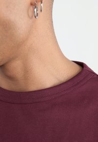 Burgunderfarget bomulls t-skjorte med rund hals, med fine sømmer langs hemmet og en jevn tekstur. Nærbilde av nakke- og skulderområdet.