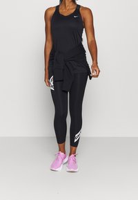 Czarny sportowy top na ramiączkach, czarne legginsy z białym logo Nike oraz różowe sneakersy z fakturowanego materiału i sznurówkami. Widoczne nogi i ręce.