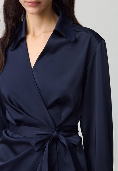 Camisa de satén azul marino con cuello, mangas largas y cintura atada. Textura suave con un brillo sutil y líneas limpias.