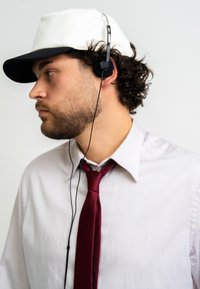 White and black baseball cap, black headphones, white striped shirt, and maroon tie. Profile view with curly hair and slight facial hair.