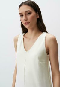 Top bianco senza maniche con scollatura a V, caratterizzato da una leggera lucentezza, una texture liscia e una piccola tasca sul lato sinistro, in un design con taglio morbido.