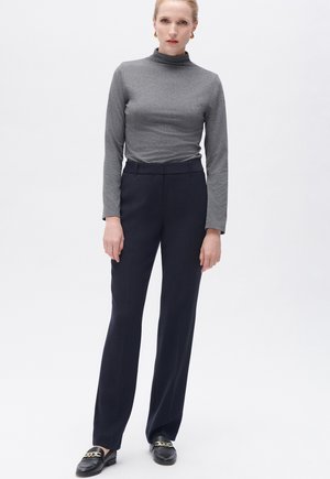 Graues, langärmliges Turtleneck-Shirt kombiniert mit navyblauen, maßgeschneiderten Hosen und schwarzen Slippern mit goldener Kettendetail. Glatte Textur, enganliegendes Design.