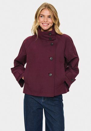 Veste bordeaux avec un col montant, fermeture à boutons et poches latérales ; fabriquée à partir d'un tissu lisse avec une coupe décontractée. Associée à un jean bleu foncé.