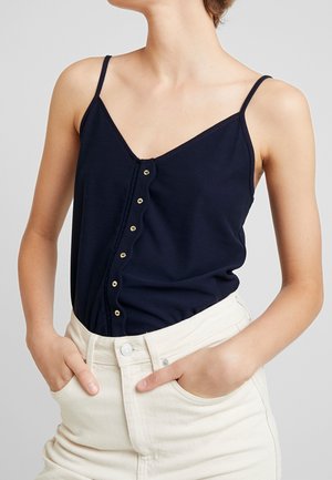 Kvinde iført en ærmeløs marineblå knap-foran-top ind tucked i højtaljede off-white bukser med hænderne i lommerne.