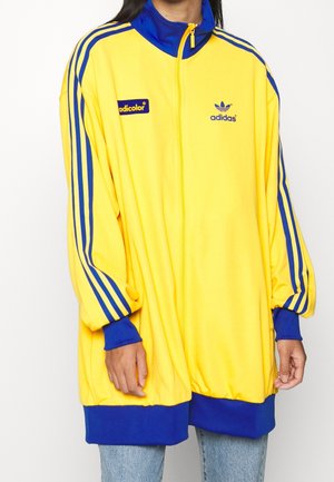 Osoba ubrana w za dużą żółtą kurtkę zapinaną na zamek marki Adidas z niebieskimi paskami na rękawach, niebieskim kołnierzem, mankietami i dolnym wykończeniem, w połączeniu z jasnoniebieskimi dżinsami.