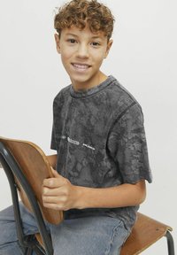 Hallitusest kujundatud hall puuvillane T-särk ümmarguse kaelusega, millel on logode detail. Mudel istub puidustToolil, hoides seljatoest.