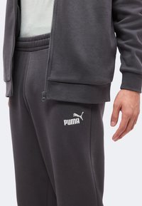 Gros plan d'une personne portant un pantalon de survêtement Puma gris foncé et une veste zippée assortie avec un logo blanc sur la cuisse gauche.