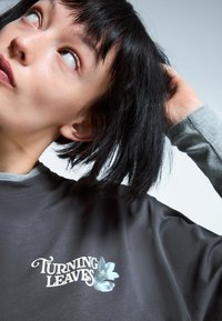 Tricou supradimensionat gri închis, cu un grafic floral și textul "ÎNTORCÂND FOLIILE". Material din bumbac moale, cu o croială lejeră.