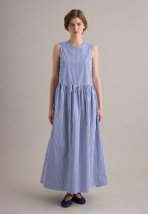 Donna in piedi che indossa un vestito lungo senza maniche a righe verticali blu e bianche e scarpe basse blu scuro su uno sfondo semplice.