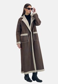 Lange bruine jas gemaakt van een imitatielederen materiaal, met een contrasterende beige shearlingkraag, manchetten en zakken.