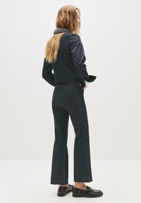 Ženska z dolgimi blond lasmi, oblečena v temno teksas jakno, usklajene hlače z visokim pasom, črne mokasine in sivo ruto, stoji v bočnem položaju.