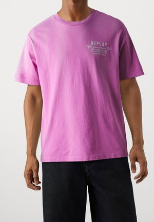 Homme portant un t-shirt rose ample avec un petit texte blanc sur la poitrine indiquant "REPLAY Get your groove on Record Store Collectable Vinyl Bought & Sold".