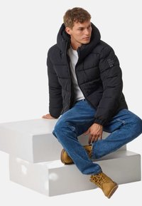 Czarna kurtka puchowa z kapturem, z zamkiem błyskawicznym i elastycznymi mankietami. Połączona z niebieskimi dżinsami i piaskowymi butami trekkingowymi na białej platformie.