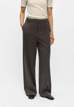 Personne portant un pantalon marron foncé taille haute et jambes larges, les mains dans les poches, avec des chaussures noires sur un fond blanc uni.