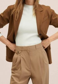 Veste tailleur marron sur un haut côtelé vert clair, associée à un pantalon beige taille haute plissé avec un détail de bouton.