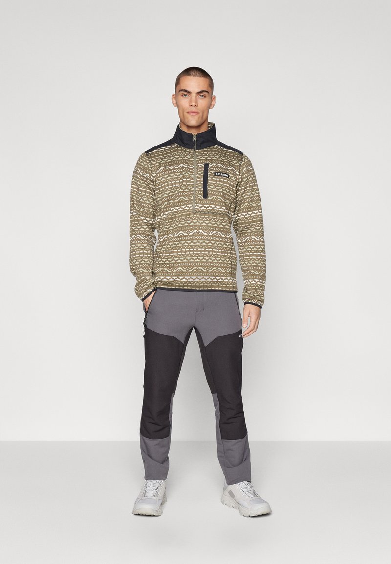 Sudadera de forro polar en verde olivo con diseño estampado, cuello con cremallera y cuello navy; combinada con pantalones grises y negros con paneles de contraste.