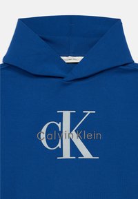 Blå bomull hettegenser med stort "CK" logo i hvitt og "CalvinKlein" i grått under. Glatt tekstur, tettsittende hette og ribbestrikket hals.