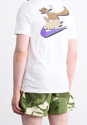 Osoba nosząca białą koszulkę z kreskówkowym kangurem i logo "NIKE", w zestawieniu z zielonymi i beżowymi, wzorzystymi szortami.