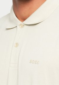 Gros plan d'une personne portant un polo beige clair avec deux boutons et le mot BOSS brodé sur la poitrine.
