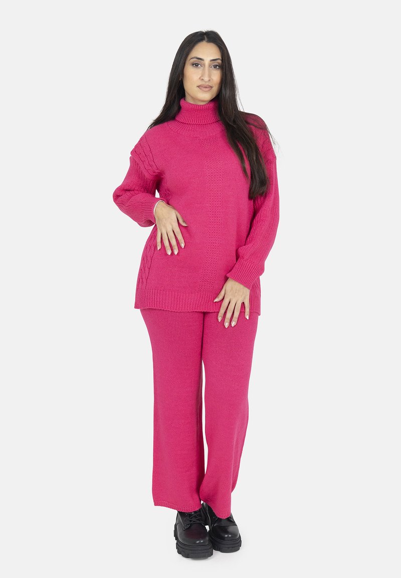 Rosa Strickpullover mit hohem Kragen und geripptem Textur, kombiniert mit passenden weiten Hosen. Schwarze klobige Stiefel runden das Outfit ab.