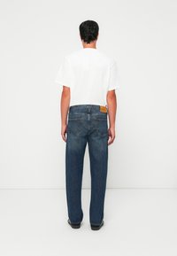 Man standing with back to camera, wearing a white short-sleeve t-shirt, blue straight-leg jeans, and black shoes against a white background.