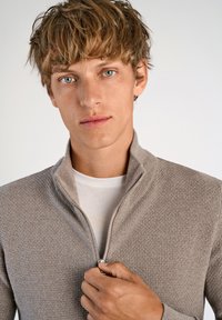 Beige strukturerad zip-up tröja med hög krage, bärs över en vit skjorta. Hand som greppar dragkedjan, vilket framhäver designens detaljer.