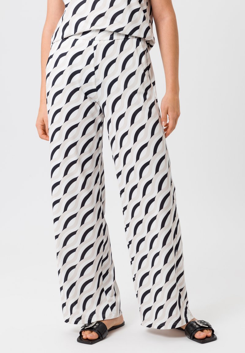 Pantalons larges blancs avec un motif ondulé en noir et gris. Tissu léger avec une texture lisse, associés à des sandales noires simples.