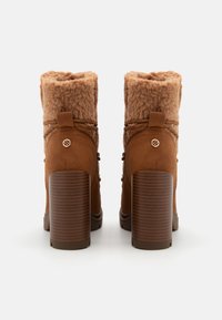 Stivali con tacco in suede marrone, con parte superiore in soffice pelliccia sintetica color tan, caratterizzati da un tacco spesso e testurizzato e dettagli hardware decorativi.