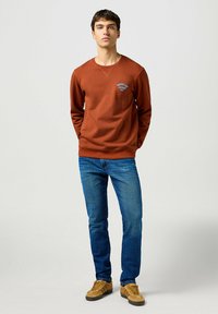Sudadera marrón con logo bordado en azul, cuello redondo, mangas largas, combinada con vaqueros azules y zapatillas color beige. Diseño simple y limpio.