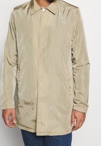 Beige lättviktsjacka med en glänsande textur, knäppning vid kragen, sidofickor och långa ärmar. Platt nederkant och minimalistisk design.