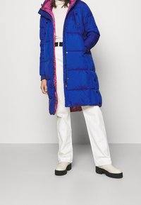 Blue puffer coat with a pink lining, featuring a hood, snap buttons, and a long silhouette. Paired with white pants and beige platform boots.