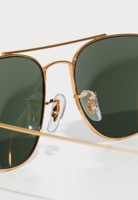 Occhiali da sole in metallo dorato con lenti verdi, design con doppio ponte, cuscinetti nasali trasparenti e logo dettagliato sulla lente.