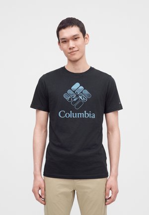 Jeune homme portant un t-shirt noir Columbia avec un logo bleu et un pantalon beige, debout devant un fond clair uni.