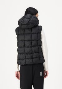 Melna pūkaina veste ar quilted dizainu un kapuci, kas izceļas ar pūkainu tekstūru un elastīgām detaļām. Valkāta virs garām piedurknēm un biksēm.