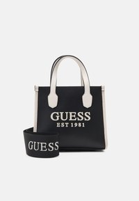 Sac à main en cuir synthétique noir avec des accents crème. Comprend un logo "GUESS EST 1981" bien visible, deux poignées supérieures et une bandoulière amovible.