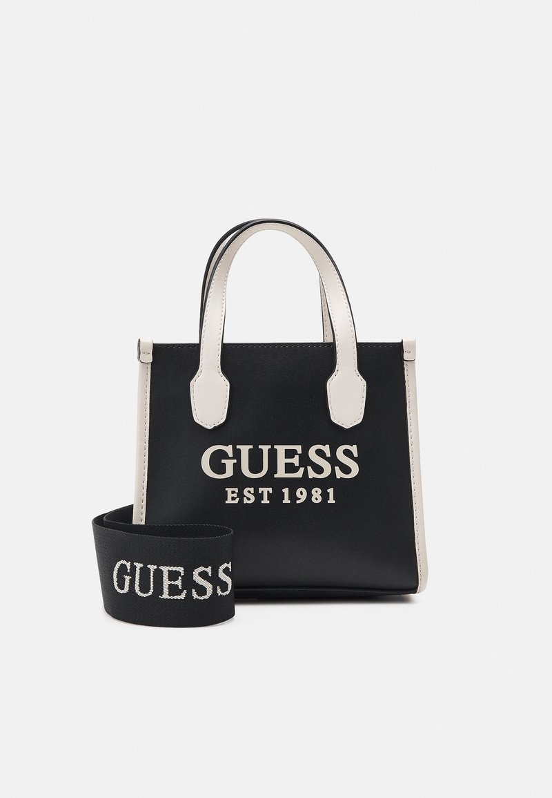 Sac à main en cuir synthétique noir avec des accents crème. Comprend un logo "GUESS EST 1981" bien visible, deux poignées supérieures et une bandoulière amovible.