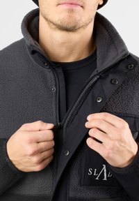 Giacca in pile nera e grigia con chiusura a zip, bottoni a pressione e tasca sul petto. Presenta una texture liscia e un design a pannelli a contrasto.
