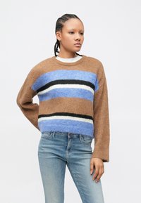 Pulover cu dungi maro și albastre, cu o textură moale, având mâneci largi și guler rotund. Asortat cu blugi albastru deschis.