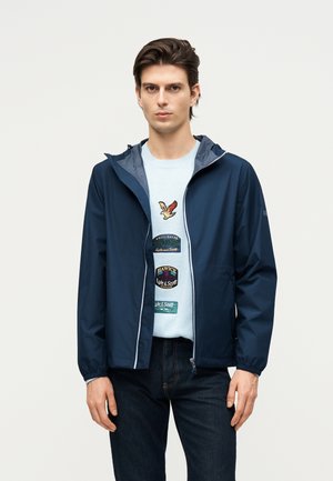 Ung mand iført en mørkeblå hættetrøje over en lyseblå sweater med fugle- og tekstpatches, kombineret med mørke jeans, stående mod en ensfarvet baggrund.